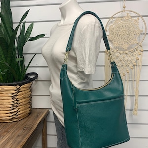 May include: A dark teal hobo bag with a pebbled texture, featuring a single shoulder strap, a top zipper closure with a leather pull, and an exterior slip pocket on the front. The bag is constructed with visible white stitching along the seams.