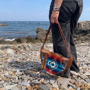 May include: A crescent-shaped shoulder bag featuring a vibrant geometric pattern in shades of blue, red, and orange. The bag includes a brown leather-like strap, a matching brown leather-like side panel, and a decorative brown tassel attached to the zipper.