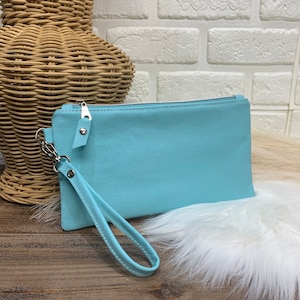 May include: A light blue leather clutch with a zipper closure and a wrist strap. The clutch is sitting on a wooden surface with a white fur rug.