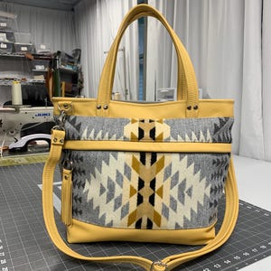 May include: A yellow and gray tote bag with a geometric pattern. The bag has a yellow leather base, handles, and shoulder strap. The main body features a gray, white, and yellow geometric design. A small tassel hangs from the strap.