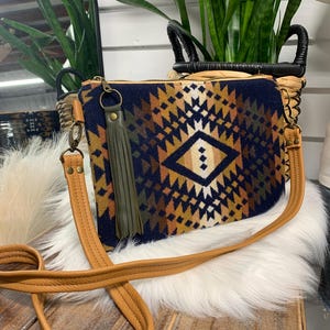 May include: A navy blue and patterned crossbody bag with a geometric design in shades of brown, tan, and white. The bag has a tan leather strap and a green tassel. The bag is sitting on a white furry surface.