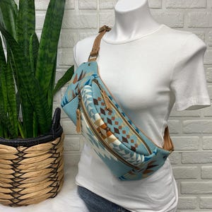 以下が含まれることがあります： 茶色、白、ターコイズの色合いの南西部のパターンが特徴の、ライトブルーのファニーパック。バッグには茶色のレザーストラップとジッパープルが付いています。ファニーパックは白いマネキンに展示されています。