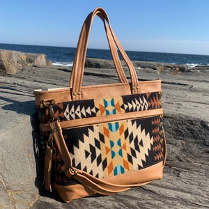 May include: A tan leather tote bag with a geometric patterned fabric panel. The bag has two top handles and a detachable shoulder strap. The geometric pattern includes black, beige, orange, and blue shapes. The bag is sitting on a rocky surface with the ocean in the background.