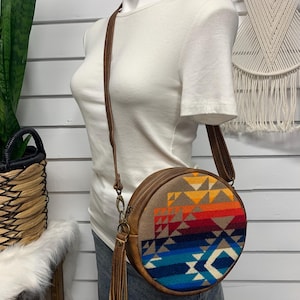 May include: Round crossbody bag with a brown leather strap and trim. The bag features a colorful geometric pattern in shades of blue, red, orange, and yellow. A brown leather tassel hangs from the zipper.