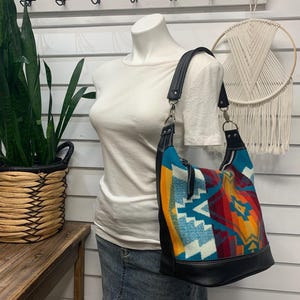 Pode incluir: Uma bolsa de ombro estilo hobo com um tecido vibrante de padrões geométricos em tons de turquesa, laranja, vermelho e amarelo, acentuada com acabamentos em couro preto, uma alça de ombro de couro preto e ferragens em tom prateado.
