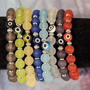 May include: A selection of beaded bracelets in shades of brown, green, yellow, blue, and red. Each bracelet features matte glass beads and decorative eye beads, connected with silver-toned metal accents. The bracelets are displayed on a black surface.