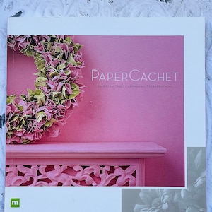 May include: A pink and white book with the title "PAPER CACHET" and the text "PAPER CRAFTING | CARDMAKING | SCRAPBOOKING". The cover features a pink paper flower wreath and a pink and white decorative shelf.