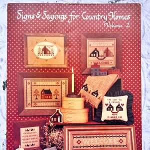 May include: A book cover with a red background and a title that reads "Signs & Sayings for Country Homes, Volume 2". The cover features several cross-stitch patterns with a country theme, including houses, trees, and welcome signs.