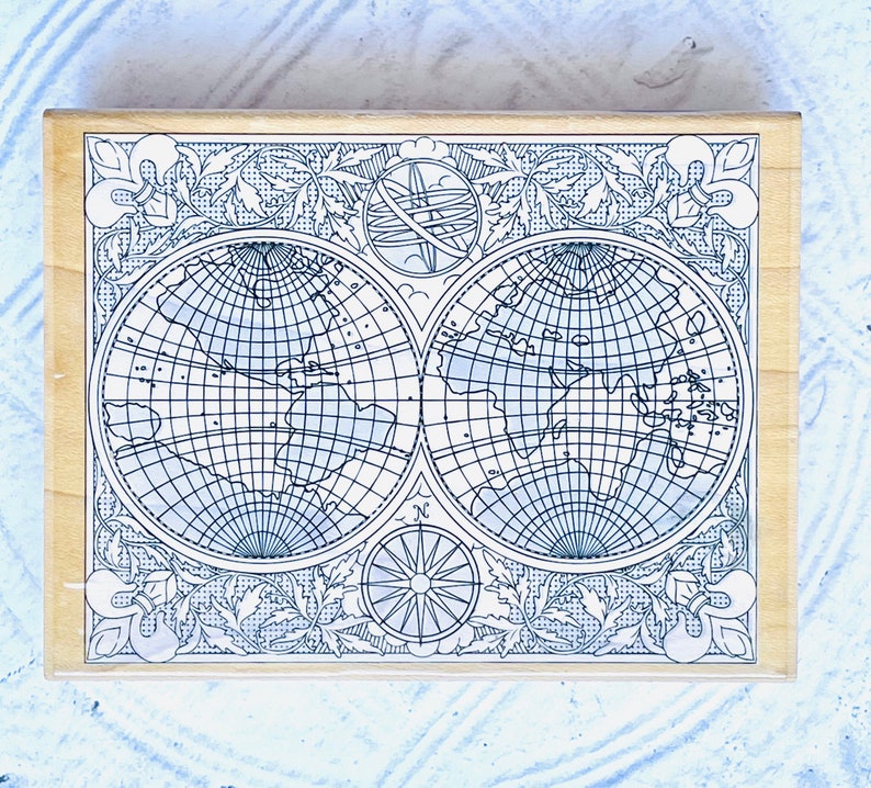 Puede incluir: Un sello de madera con un dise&ntilde;o vintage que presenta dos mapas del mundo, una br&uacute;jula y detalles florales. Los mapas est&aacute;n delineados en negro y tienen un patr&oacute;n de cuadr&iacute;cula.