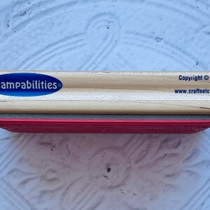 Puede incluir: Un sello de madera con una superficie de goma roja. El sello tiene una etiqueta ovalada azul que dice "Stampabilities" con un s&iacute;mbolo de marca registrada. La parte inferior del sello tiene el texto "Copyright &copy; 2002 www.craftsetc.com".