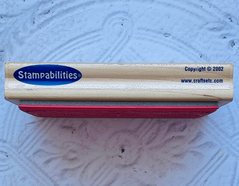 Puede incluir: Un sello de madera con una superficie de goma roja. El sello tiene una etiqueta ovalada azul que dice "Stampabilities" con un s&iacute;mbolo de marca registrada. La parte inferior del sello tiene el texto "Copyright &copy; 2002 www.craftsetc.com".