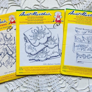 May include: Three vintage Aunt Martha's Hot Iron Transfer packets. The packets are yellow with red and blue text and illustrations. Designs include embroidery patterns for pillowcases, a bedroom ensemble, and kittens.