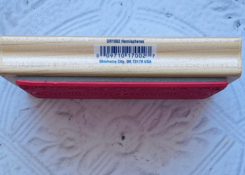 Puede incluir: Un sello de madera con una almohadilla de goma roja. El sello est&aacute; etiquetado como "DR1002 Hemispheres" y tiene un c&oacute;digo de barras con el n&uacute;mero "0 09710 17002 7". El texto "Oklahoma City, OK 73179 USA" tambi&eacute;n est&aacute; impreso en el sello.