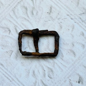 May include: A rusty metal buckle with two rectangular openings. The buckle is a dark brown color and has a worn, aged appearance.
