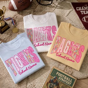 May include: Three t-shirts in light blue, white, and pale yellow featuring pink patterned block letters that spell TIGERS, HAWKS, and EAGLES with the text School Name below each. The shirts are arranged with a football, a trophy, and a book.