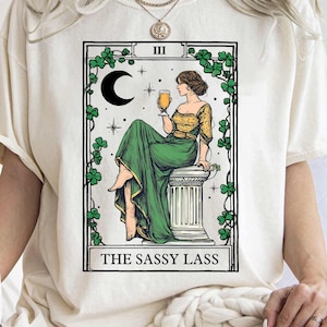 May include: Cream-colored t-shirt featuring a tarot card design. The card depicts a woman in a green dress holding a drink, with the text "THE SASSY LASS" at the bottom. The design includes a crescent moon, stars, and decorative ivy.