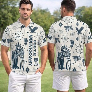 May include: Cream-colored polo shirt with a navy blue print featuring the Statue of Liberty, American flags, eagles, and fireworks. The shirt has the text "1776-2026 250th Anniversary". The model is wearing white shorts.