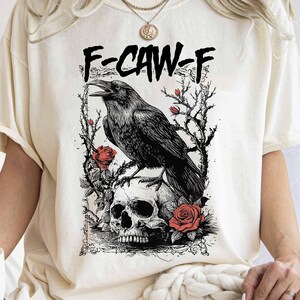 Bring dark elegance and bold funny symbolism to your wardrobe with this F-Caw-F gothic raven t-shirt. Designed in a vintage hand-drawn sketching style, the illustration features a funny crow perched on thorny climbing roses, looming over a detailed human skull. Heavy black line art, engraved shading, and distressed ink textures create a dramatic tarot-inspired design. Deep crimson roses accent the composition, adding a striking contrast against the bold black artwork.