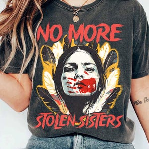May include: A dark grey t-shirt with a graphic print of a Native American woman with her mouth covered in red paint. The text "NO MORE STOLEN SISTERS" is printed on the shirt.