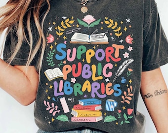 May include: Dark grey t-shirt with the text "Support Public Libraries" in colourful letters. The design includes illustrations of books, flowers, glasses, and a feather. The shirt is a vintage style.