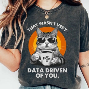 May include: Dark gray t-shirt featuring a cat wearing sunglasses and holding a coffee mug. The shirt has the text "THAT WASN'T VERY DATA DRIVEN OF YOU." The cat is set against an orange and yellow circular background.
