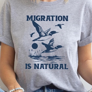 May include: A light gray t-shirt with the words "MIGRATION IS NATURAL" in navy blue. The shirt features a graphic of flying birds over water, with a sun and reeds.