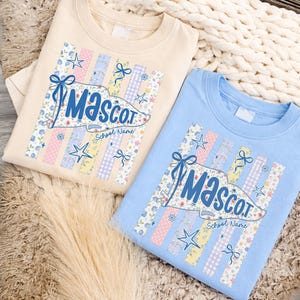 May include: Two crew neck sweatshirts, one beige and one light blue, each with a floral and star design. The design includes the word "Mascot" in blue script, with space for a school name.