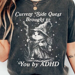 Celebrate neurodivergent humor with this Current Side Quest Brought to You by ADHD graphic t-shirt. This shirt is made for those who embrace mental health awareness, side quests, and a sense of humor about everyday distractions. Ideal for ADHDers, gamers, RPG lovers, and anyone who lives life one side quest at a time.
