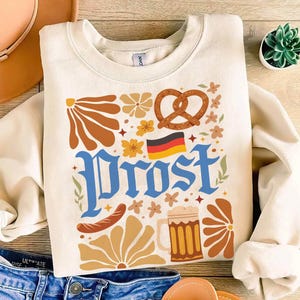 Celebrate Oktoberfest in true Bavarian style with this Prost Oktoberfest Shirt! The design features bold retro lettering, a frothy beer mug, pretzel, baguette, and German flag surrounded by floral accents – a perfect nod to German beer culture and festival vibes. Whether you’re heading to an Oktoberfest party, brewery night, or just love craft beer, this cozy unisex tee makes the ultimate drinking shirt.