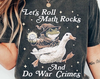 May include: Dark gray t-shirt with the text "Let's Roll Math Rocks And Do War Crimes." The graphic features a frog in a wizard hat holding a die, riding a white goose. The shirt is a vintage style.