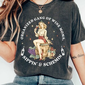May include: Dark grey t-shirt with a circular graphic of a woman with wine, seated on a barrel. Text reads "ORGANIZED GANG OF WINE MOMS, SIPPIN' & SCHEMIN". Two wine bottles flank the graphic.