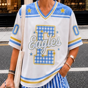 May include: A white V-neck jersey with blue and gold stripes, featuring a large blue gingham letter E with the word Eagles in script, surrounded by small gold stars. The jersey is paired with blue and white vertical striped shorts.