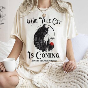 May include: Cream-colored t-shirt with a black graphic of a cat silhouette over a cityscape, a red sun, and the text "The Yule Cat Is Coming. Beware the Jolakottrinn."