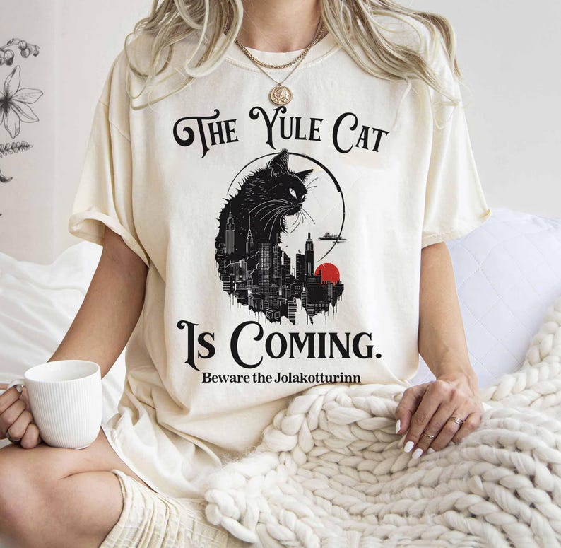 May include: Cream-colored t-shirt with a black graphic of a cat silhouette over a cityscape, a red sun, and the text "The Yule Cat Is Coming. Beware the Jolakottrinn."