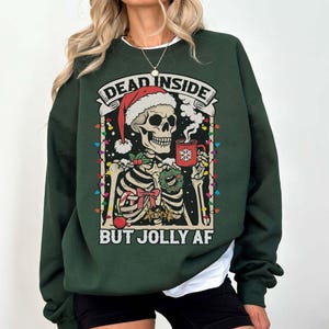 May include: Green Christmas sweater with a skeleton wearing a Santa hat and holding a mug of hot cocoa. The sweater has the text "Dead Inside But Jolly AF" printed on it.