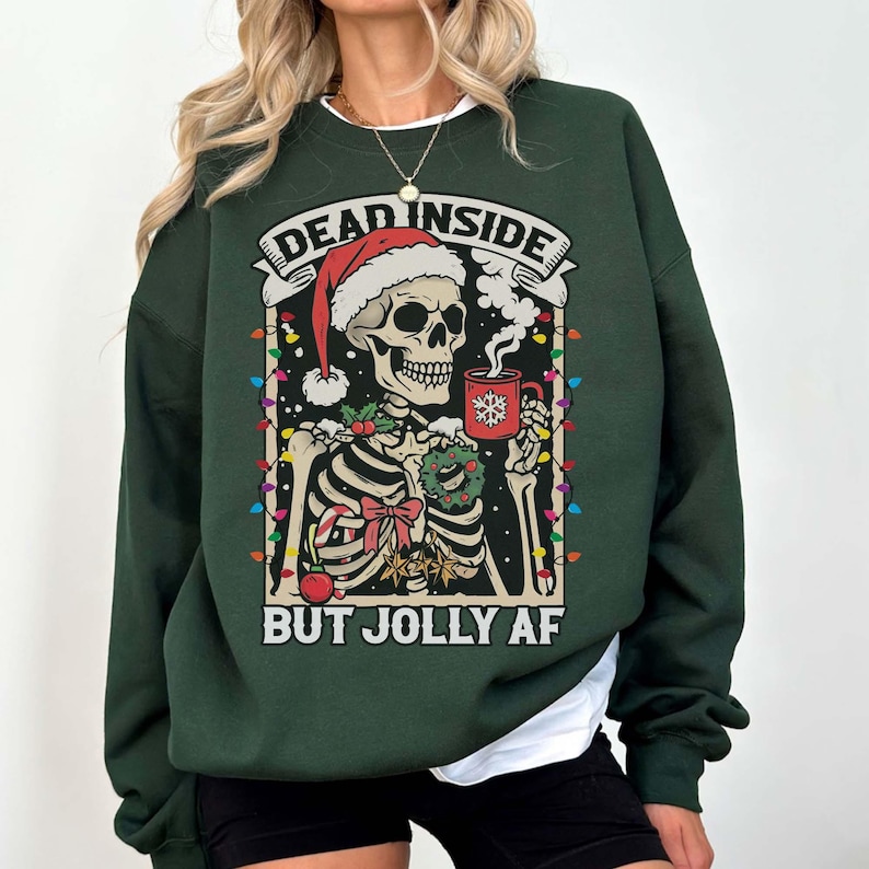 May include: Green Christmas sweater with a skeleton wearing a Santa hat and holding a mug of hot cocoa. The sweater has the text "Dead Inside But Jolly AF" printed on it.