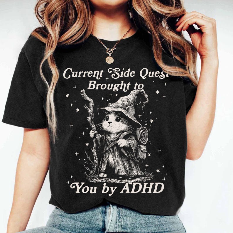 Pode incluir: T-shirt preta com um desenho branco de um gato mago segurando uma bengala e usando um chap&eacute;u. O texto diz "Current Side Quest Brought to You by ADHD". O design se destaca em um fundo escuro, criando um contraste marcante.