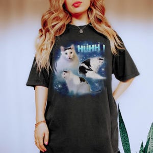 May include: Black t-shirt with a graphic of three cats set against a space-themed backdrop and the word "HUHH!". The cats are white and black. The t-shirt has a relaxed, oversized fit.