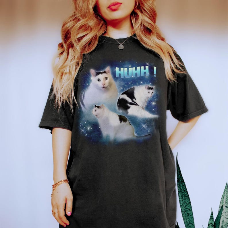 May include: Black t-shirt with a graphic of three cats set against a space-themed backdrop and the word "HUHH!". The cats are white and black. The t-shirt has a relaxed, oversized fit.