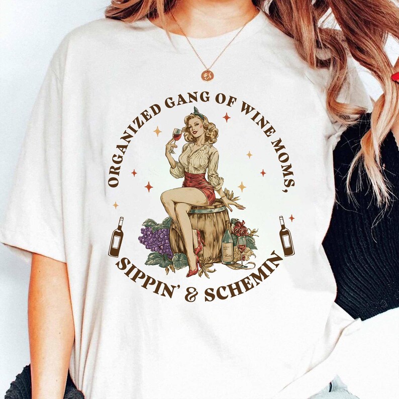 May include: Cream-coloured t-shirt with a retro illustration. It depicts a woman on a barrel with a wine glass, grapes, and bottles. The text reads "ORGANIZED GANG OF WINE MOMS, SIPPIN' & SCHEMIN'" in a circular arrangement around the image.