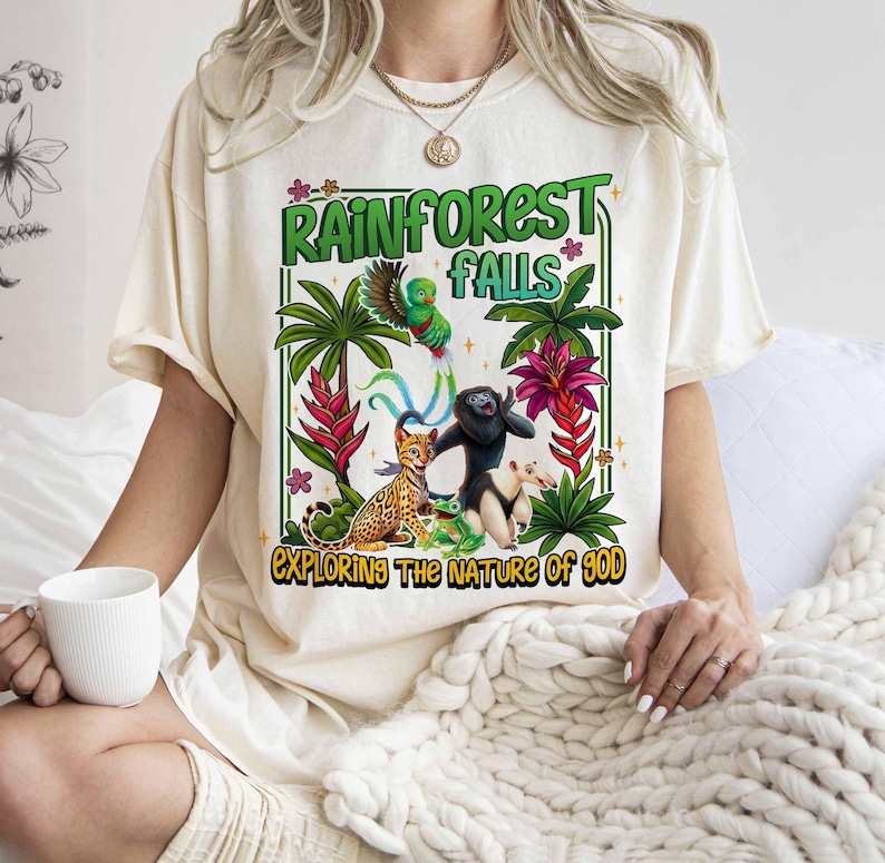 May include: Cream-colored t-shirt featuring a vibrant rainforest design. The graphic includes the text "Rainforest Falls" and "Exploring the Nature of God", along with illustrations of a parrot, leopard, monkey, and other animals.