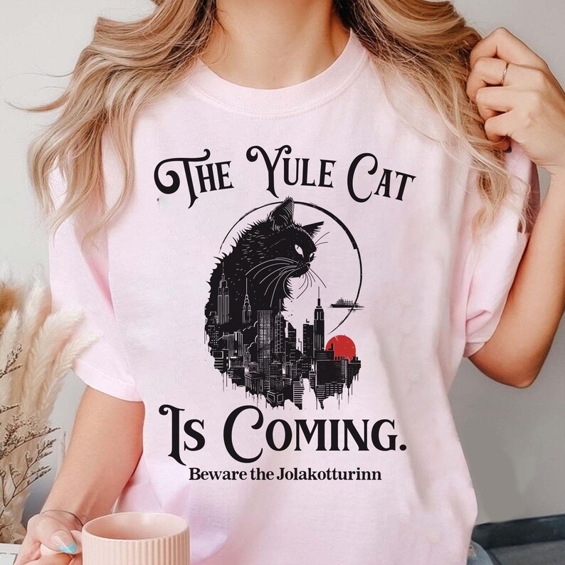 May include: Light pink t-shirt with the text "The Yule Cat Is Coming." A black cat silhouette is set against a cityscape and a red sun. The phrase "Beware the Jolakottturinn" is below.