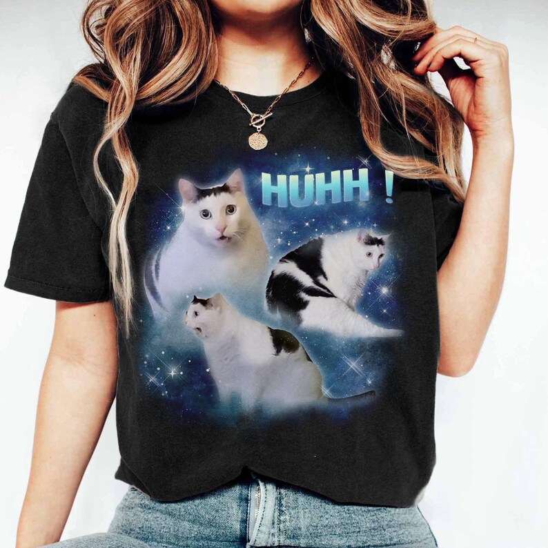 May include: Black t-shirt with a graphic of three cats and the text "HUHH!". The cats are white with black patches, set against a blue, starry backdrop. A humorous design, suitable for casual wear.