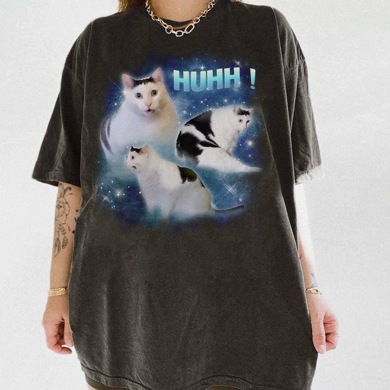 May include: Dark grey t-shirt with a graphic of three cats against a space-themed backdrop and the word "HUHH!". The cats are white and black, set against a blue and white starry sky.