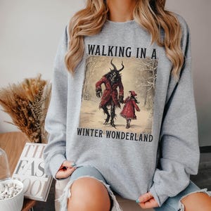 May include: A light gray sweatshirt featuring a winter scene with the text "WALKING IN A WINTER WONDERLAND." The graphic depicts a horned figure in a red coat and a child in a red dress walking in a snowy forest.