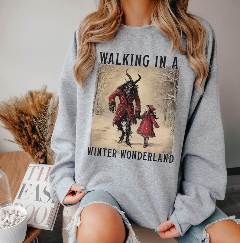 May include: A light gray sweatshirt featuring a winter scene with the text "WALKING IN A WINTER WONDERLAND." The graphic depicts a horned figure in a red coat and a child in a red dress walking in a snowy forest.