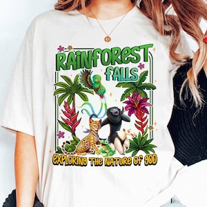 May include: White t-shirt with a colorful rainforest graphic. The design features a green parrot, a black monkey, a spotted cat, a frog, and an anteater, with the text "RAINFOREST FALLS" and "EXPLORING THE NATURE OF GOD".