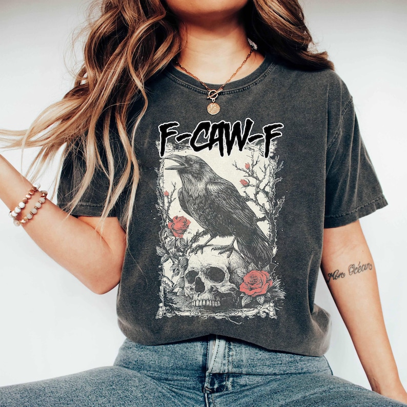 Puede incluir: Camiseta gris oscuro con un dise&ntilde;o de cuervo negro posado sobre una calavera, rodeado de rosas y ramas. El texto "F-CAW-F" est&aacute; encima del gr&aacute;fico. La camiseta tiene un estilo vintage, probablemente hecha de algod&oacute;n o una mezcla de algod&oacute;n.