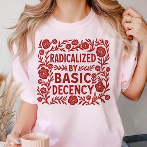 May include: Light pink t-shirt with the text "RADICALIZED BY BASIC DECENCY" in a red floral design. The design features flowers and leaves around the text. The shirt is a crew neck.