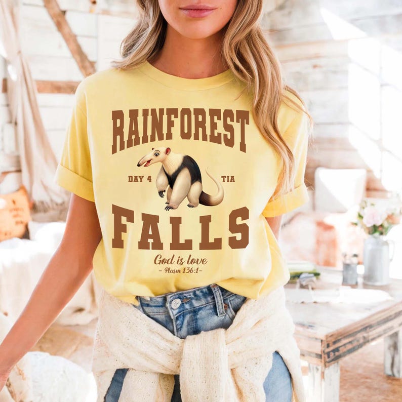 May include: Yellow t-shirt with the words "RAINFOREST FALLS" in brown block letters. A cartoon anteater is centered below the word "RAINFOREST". The text "God is love - Psalm 136:1-" is printed at the bottom of the shirt.